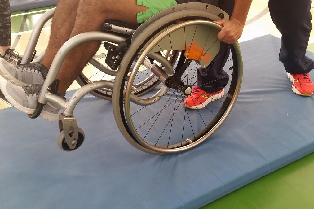 zur Vergrößerungsansicht des Bildes: Eine Person übt das Balanciern im Rollstuhl auf einer weichen Matte, eine zweite Person hilft.