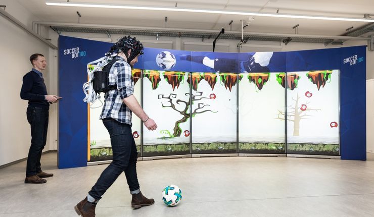 Eine Person läuft durch das Forschungslabor der Sportwissenschaftlichen Fakultät. Sie trägt Senstoren zur Infrarotspektroskopie auf dem Kopf. Vor ihren Füßen liegt ein Fußball.