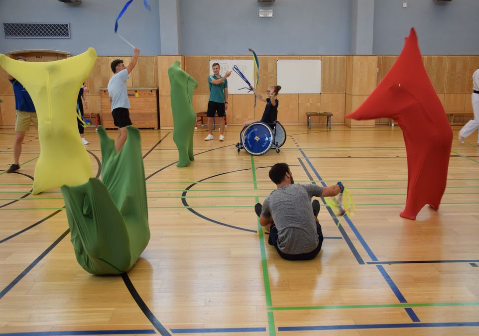 zur Vergrößerungsansicht des Bildes: In einer Sporthalle sind viele Studierende, die sich mit unterschiedlichem Material bewegen, z.B. bunte Tanzsäcke, Kiwidos und Bänder. 