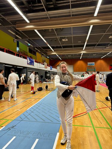 Eine Person im Fechtanzug steht zentral in einer Sporthalle und präsentiert eine T-Shirt.