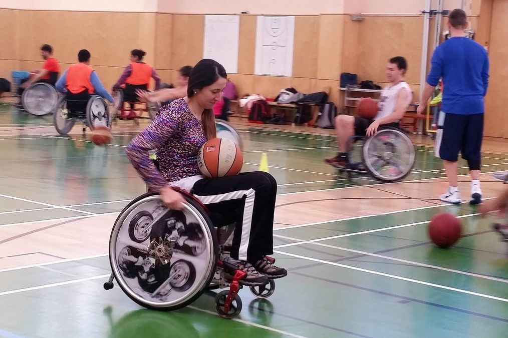 Rolstuhlbasketball - Eine Frau sitzt im Rollstuhl, führt den Ball auf dem Schoß. Andere spielen im Hintergrund.
