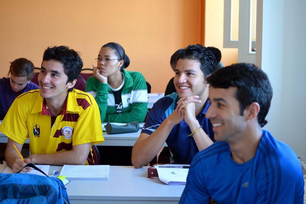 zur Vergrößerungsansicht des Bildes: 5 Studenten sitzen im Seminarraum an ihren Tischen und verfolgen lachend den Unterricht