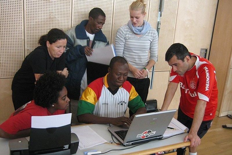 Vier Personen sitzen an einem Tisch neben den Basketballfeld und verfolgen eine Szene am Computer. Die Lehrerin erklärt.