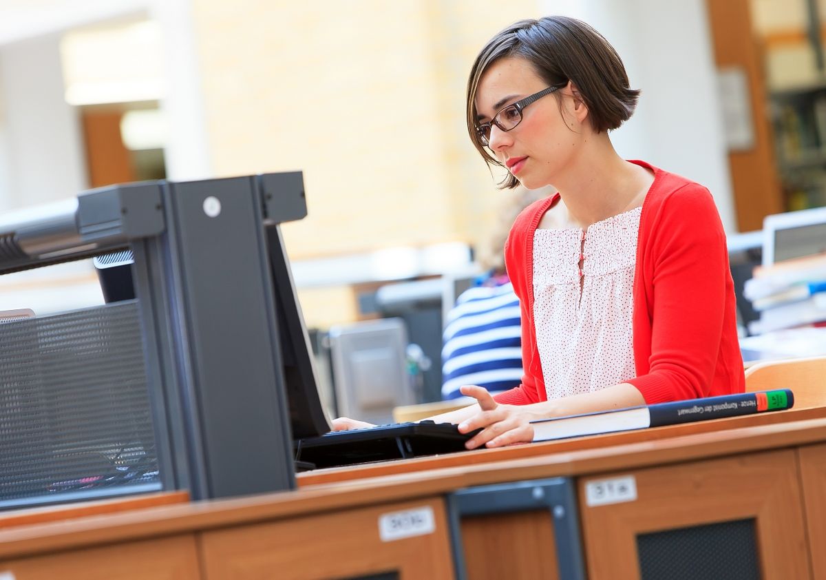zur Vergrößerungsansicht des Bildes: Foto: Eine junge Frau sitzt in der Universitätsbibliothek am Laptop und arbeitet.