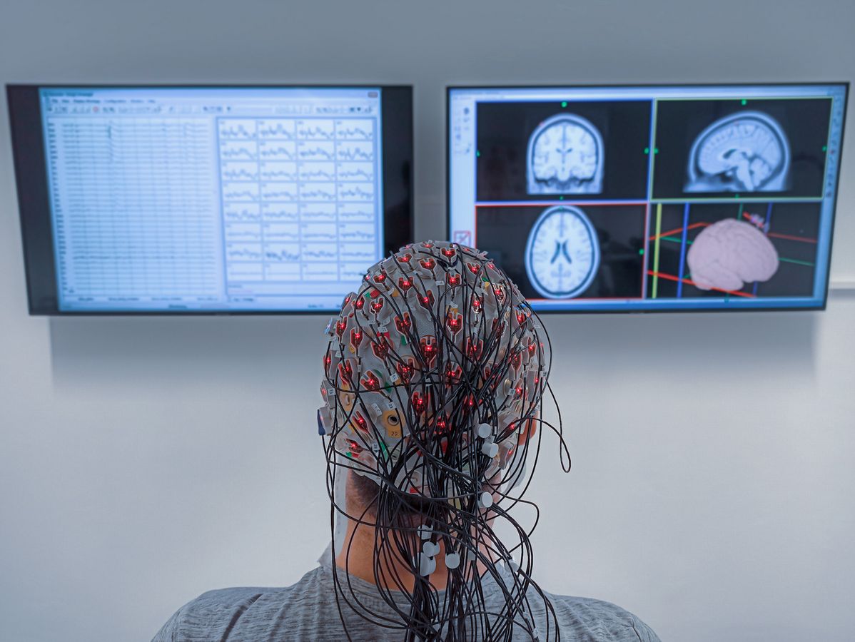 zur Vergrößerungsansicht des Bildes: Ein Proband trägt eine EEG-Haube und im Hintergrund sieht man auf zwei Monitoren die Auswertung