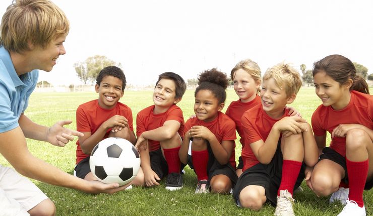 ein Fußballtrainer zeigt sechs Kindern in roten Trikots einen Fußball