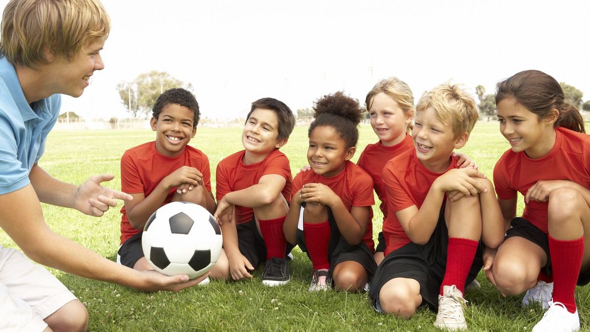 zur Vergrößerungsansicht des Bildes: ein Fußballtrainer zeigt sechs Kindern in roten Trikots einen Fußball