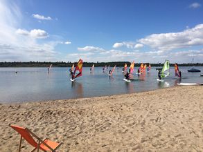 Blick vom Strand auf den Schladitzer See mit einer Gruppe von Studierenden auf Windsurfboards