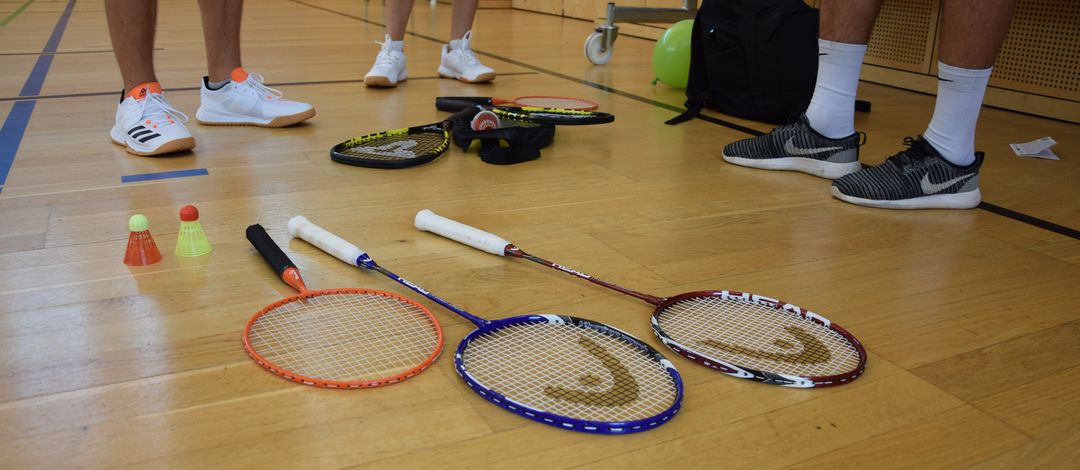 Drei Badmintonschläger mit verschiedenen Schlägerlängen und zwei Federbälle liegen auf dem Boden. Dahinter sieht man die Füße von drei Studierenden. 