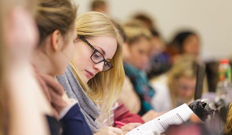 Im Blick ist eine Sitzreihe von der Seite mit Fokus auf eine Studentin, die gerade etwas aufschreibt, Foto: Christian Hüller