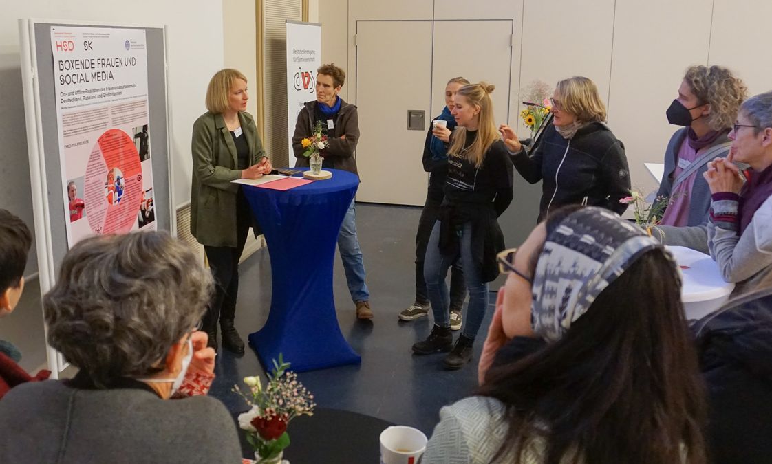 zur Vergrößerungsansicht des Bildes: Auf dem Bild sind verschiedene Personen zu sehen, die vor einem Poster diskutieren.