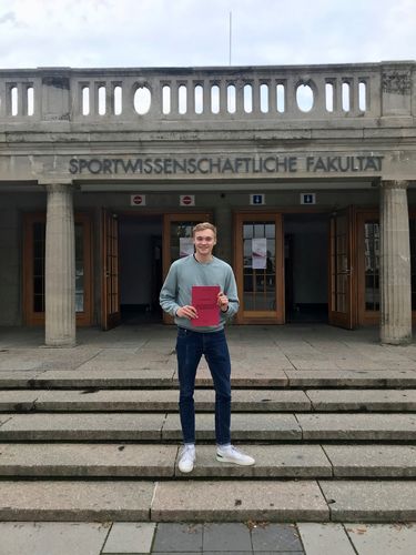 Das Bild zeigt Herrn Torben Streich mit seiner Bachelorarbeit vor dem Eingang der Sportwissenschaftlichen Fakultät