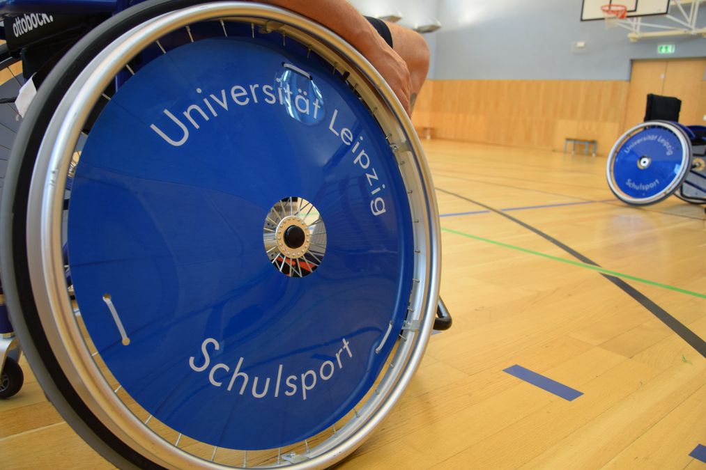 In einer Sporthalle stehen zwei Sportrollstühle. Auf dem Radschutz des vorderen steht "Universität Leipzig Schulsport" geschrieben. 