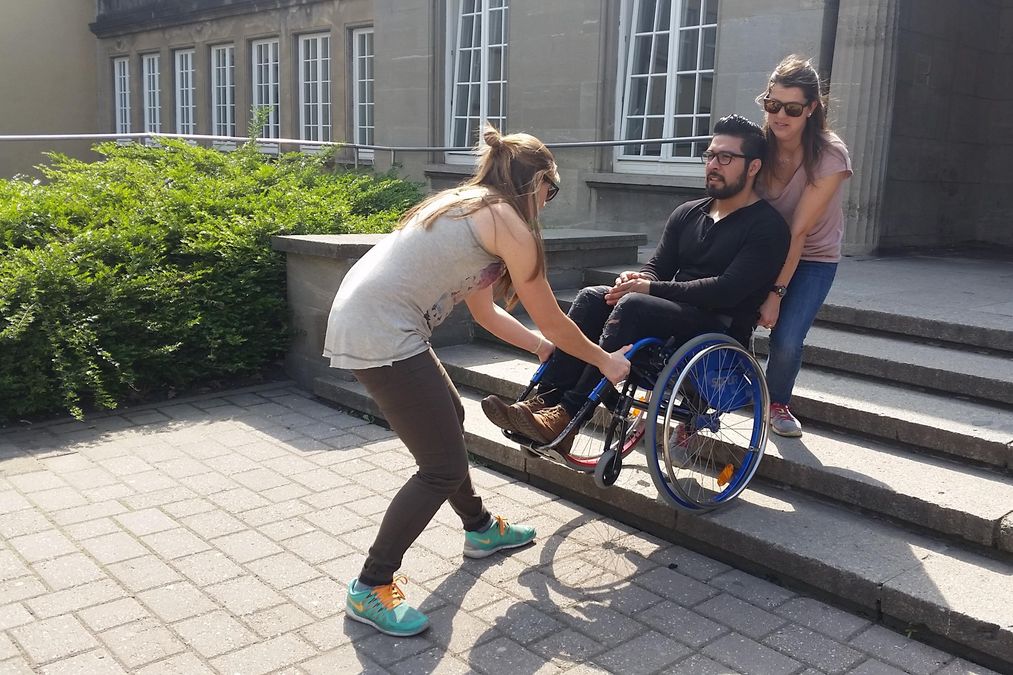 enlarge the image: Zwei Teilnehmerinnen tragen einen Mann im Rollstuhl die Treppen hinunter.