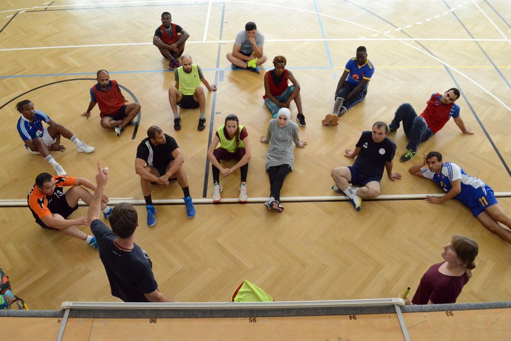 Eine Handballgruppe sitzt gemeinsam am Boden und lässt sich an der Tafel etwas erklären.