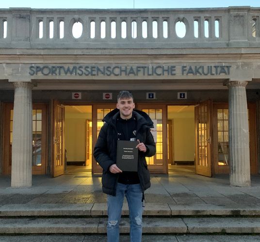 Das Foto zeigt Tobias Herrmann vor dem Eingang der Sportwissenschaftlichen Fakultät mit seiner Bachelorarbeit.