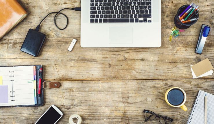 Farbfoto: Ausschnitt Holztisch mit Laptoptastatur mit angeschlossener Fstplatte, rechts daneben Stiftehalter mit Stiften darunter Tacker kleine Notizzettle, eine Tasse Kaffe, ein Block mit Stift und eine Brille. Auf der linken Seite eine Rolle Tesafilm, Handy und ein Taschenkalender.
