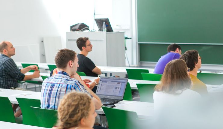 Studierende sitzen im Hörsaal an Tischen verteilt und folgen einer Vorlesung