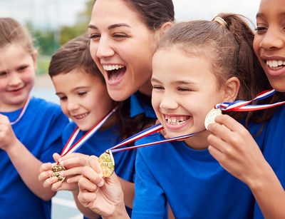 Teaserfoto für das Präventionsprojekt 2Steps4Health. Das Foto zeigt 4 Kinder und eine erwachsene Person, alle tragen blaue Shirts und die Kinder zeigen lachend ihre Medaillen in die Kamera, die sie umhängen haben.