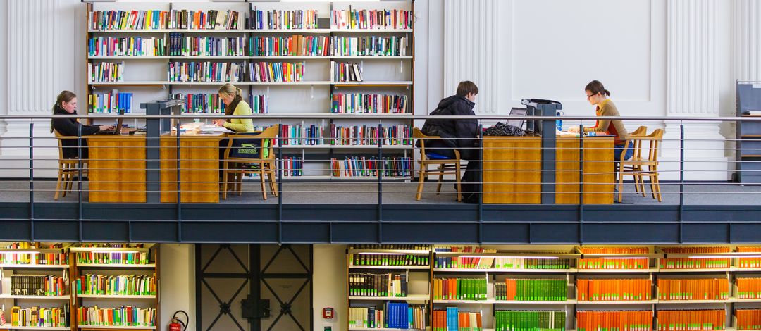 Zu sehen sind Studierende die in der Albertina-Bibliothek vor vollen Bücherregalen lernen. 
