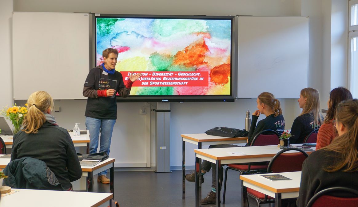 zur Vergrößerungsansicht des Bildes: Heike Tieman steht vor den Teilnehmenden der Nachwuchstagung. Im Hintergrund ist auf einem Smartboard das Tagung-Cover zu sehen.